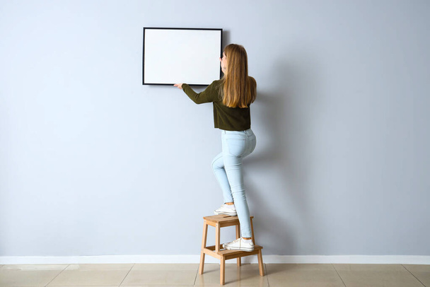 Молода жінка висить порожня рамка на світлій стіні вдома
 - Фото, зображення