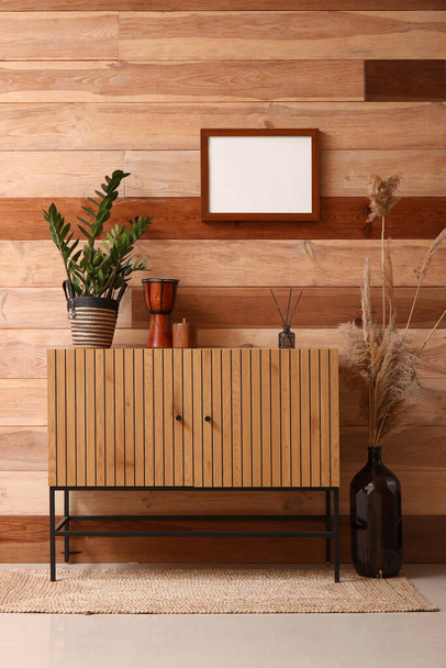 Ladekast met kamerplant, trommel en riet diffuser in de buurt van houten wand in de kamer - Foto, afbeelding