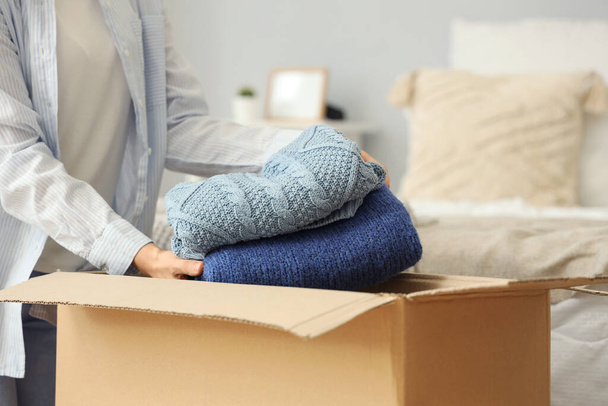Молодая женщина упаковывает одежду в картонную коробку в спальне, крупным планом - Фото, изображение