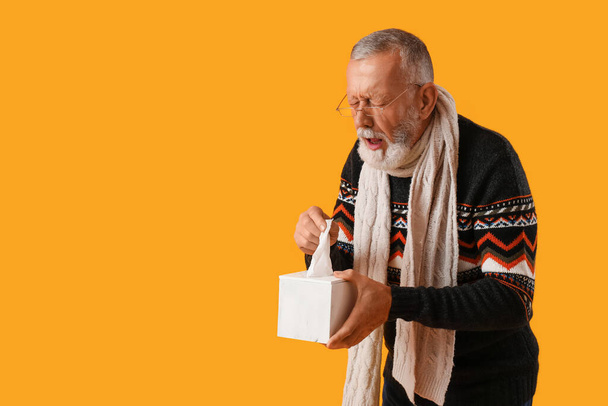 Hombre maduro enfermo con caja de tejido estornudando sobre fondo amarillo - Foto, Imagen
