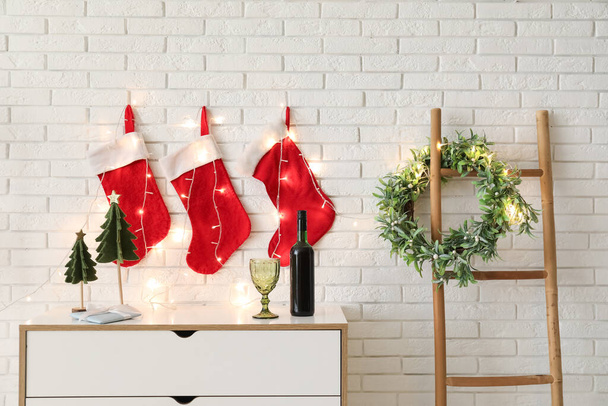 Komoda s dekorativní jedlí, lahví vína a vánočními ponožkami zavěšenými na zdi z bílých cihel - Fotografie, Obrázek