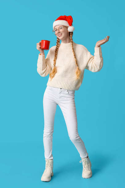 Mooie jonge gelukkige vrouw in Santa hoed met kopje thee op blauwe achtergrond - Foto, afbeelding