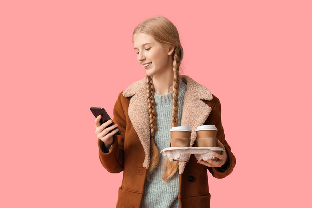 Jonge gelukkige vrouw met papieren kopjes thee en mobiele telefoon op roze achtergrond - Foto, afbeelding