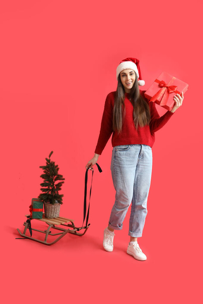 Νεαρή γυναίκα σε Σάντα καπέλο με χριστουγεννιάτικα δώρα, έλατο και έλκηθρο σε κόκκινο φόντο - Φωτογραφία, εικόνα