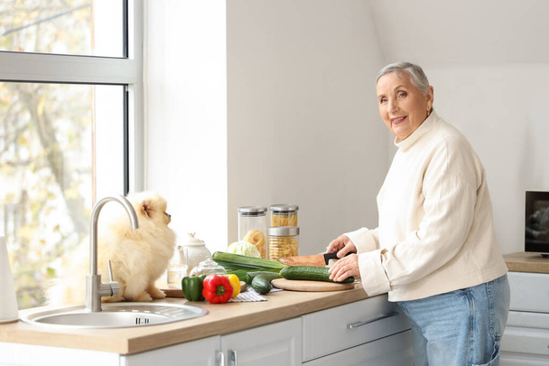 Senior vrouw met Pomeranian hond snijden groenten in de keuken - Foto, afbeelding