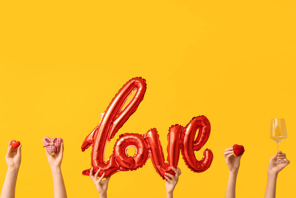 Manos femeninas con globos aéreos en forma de palabra AMOR, corazones decorativos y vidrio sobre fondo amarillo. Celebración de San Valentín - Foto, imagen
