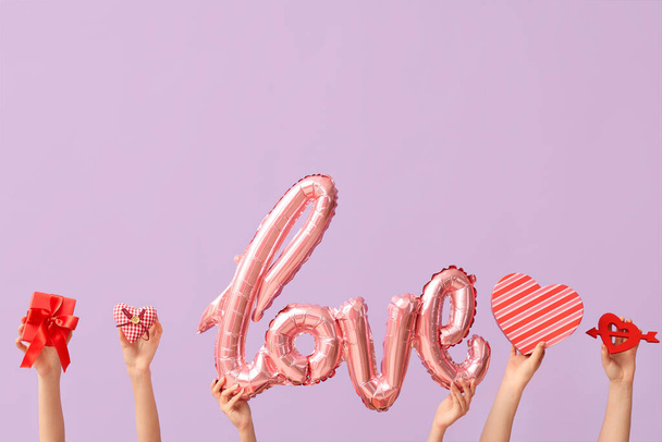 Vrouwelijke handen met luchtballonnen in de vorm van woord LOVE, decoratieve harten en geschenkdozen op lila achtergrond. Valentijnsdag - Foto, afbeelding