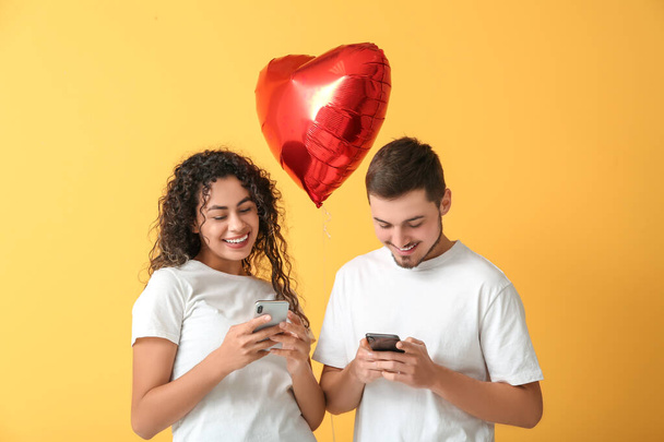 Young couple with mobile phones and air balloon in shape of heart for Valentine's day on yellow background - Photo, Image