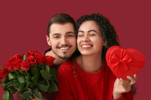 Pareja joven con flores de rosas y regalo para el día de San Valentín sobre fondo rojo - Foto, imagen