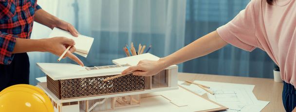 スマートアーキテクトエンジニアとインテリアデザイナーが協力し,ブループリントと建築機器の会議テーブルにルーラーを使用して家モデルを測定します. チームワークコンセプト。 バージョニング. - 写真・画像