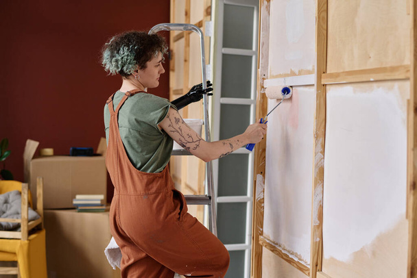 Vue arrière de la jeune femme avec un bras prothétique debout sur l'échelle et le mur de peinture dans l'appartement - Photo, image