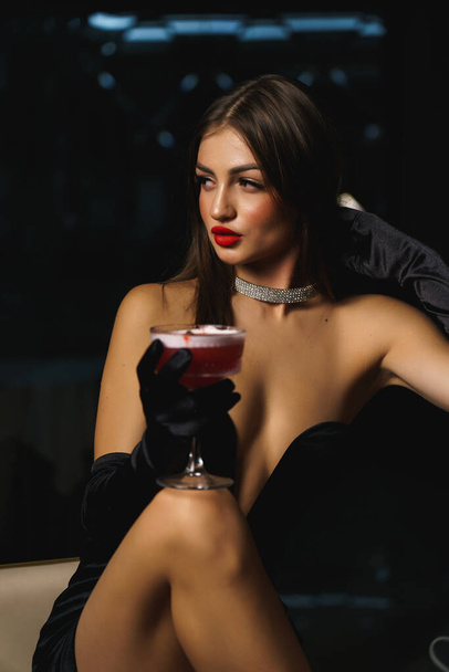 Sexy Frau im schwarzen Kleid mit Handschuhen und roten Lippen hält einen Cocktail in der Hand. - Foto, Bild