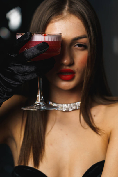 Seksikäs nainen mustassa mekossa, jossa on käsineet ja punaiset huulet, pitää cocktailin. - Valokuva, kuva