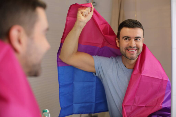 portret van knappe jongeman met biseksuele vlag voor spiegel in badkamer - Foto, afbeelding