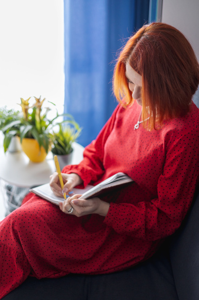 młoda kobieta z rudymi włosami siedzi na kanapie i robi notatki w notatniku - Zdjęcie, obraz