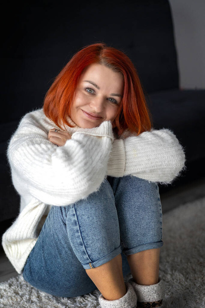 νεαρή γυναίκα με φωτεινά κόκκινα μαλλιά κάθεται στο πάτωμα στο σπίτι σε ένα λευκό ζεστό πουλόβερ - Φωτογραφία, εικόνα