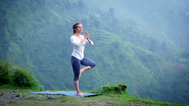 Nő gyakorlatok jóga asana Vrikshasana fa pózol a Himalája hegység szabadban egyensúlyt. Himachal Pradesh, India. Panoráma - Felvétel, videó