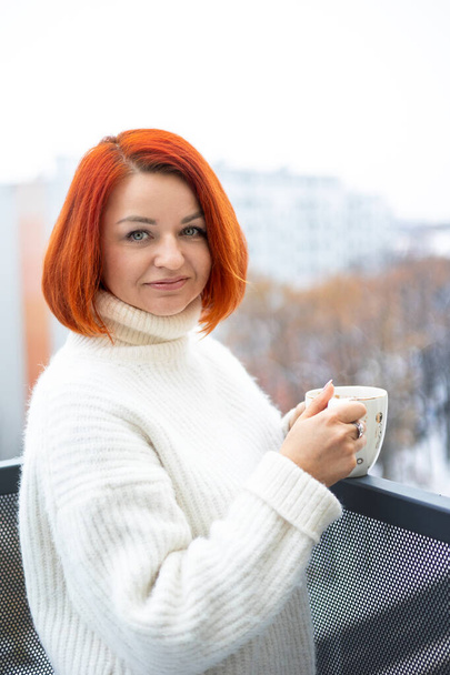 Νεαρή γυναίκα με κόκκινα μαλλιά πίνει καφέ σε μπαλκόνι με κρύο καιρό - Φωτογραφία, εικόνα