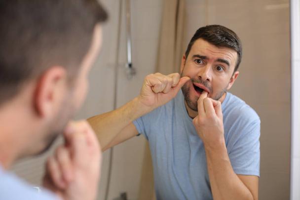 portrait of handsome young man using floss to brush teeth in front of mirror in bathroom - Photo, Image