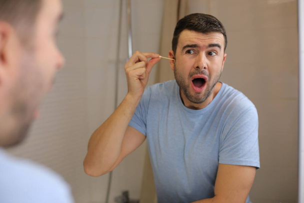 портрет красивого молодого человека, чистящего уши перед зеркалом в ванной комнате - Фото, изображение