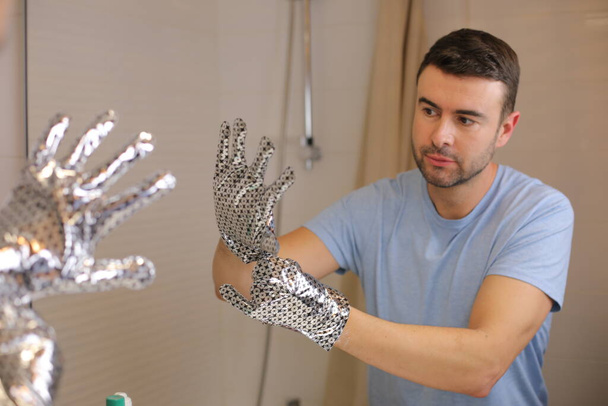πορτρέτο του όμορφος νεαρός άνδρας με ασημένια γυαλιστερά γάντια μπροστά από τον καθρέφτη στο μπάνιο - Φωτογραφία, εικόνα