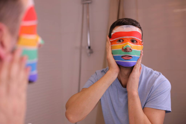 portrét pohledného mladého muže s obličejovou maskou v barvách vlajky Igbtqa před zrcadlem v koupelně - Fotografie, Obrázek