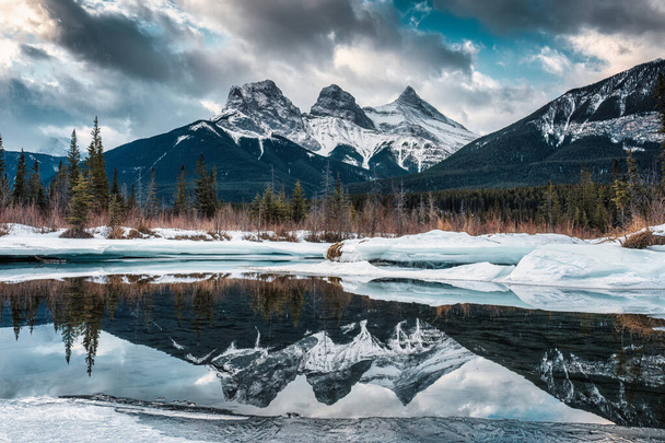 Hermosa vista de las montañas Three Sisters con nieve cubierta en el reflejo del río arco congelado en la mañana del invierno en Canmore, Alberta, Canadá - Foto, imagen