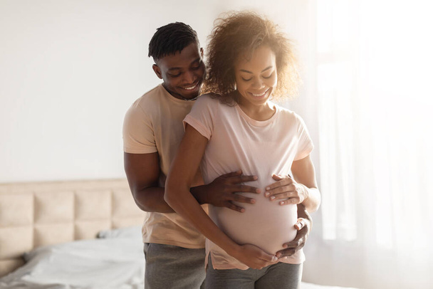 Mutlu siyahi çiftin sıcak kucaklaşmalarında, kocalarının elleri evde rahat yatak odalarında hamile eşlerinin karnına hafifçe dokunuyor. Ebeveynliğin sevgisi ve beklentisi - Fotoğraf, Görsel