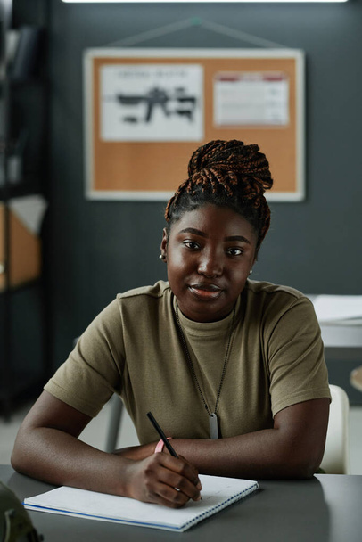 Jonge serieuze Afro-Amerikaanse vrouw met pen in de hand kijken naar de camera terwijl zitten aan het bureau en het maken van lezing notities - Foto, afbeelding