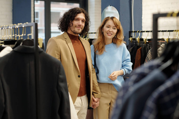 Mladý láskyplný pár v klidné luxusní oblečení stojící v butiku a při pohledu na novou sezónní kolekci módního oblečení - Fotografie, Obrázek
