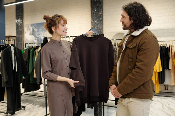 若い笑顔の女性は,男性のための新しいスウェットシャツでハンガーを保持しながら,彼女の前に立っている暖かいジャケットで顧客にそれを示しています - 写真・画像