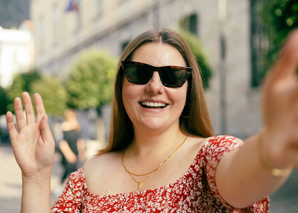 Nuori kaunis tyttö tervehtii. Valokuva rento viehättävä positiivinen nainen pidä kamera ja tehdä selfie kesällä ulkona kaupungin keskustassa ulkona. Elämäntapa turisti matka loma käsite. - Valokuva, kuva
