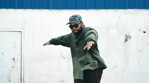 Hipster tanzender B-Boy tritt auf Straße mit blauer Wand. Asiatische Breakdancer üben Streetdance, während sie die Arme ausstrecken. Hip-Hop-Choreographen führen energiegeladene Tänze auf. Outdoor-Sport 2024. Vorfreude. - Foto, Bild