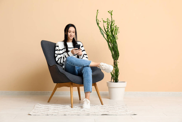 Schöne junge Frau mit Handy sitzt auf bequemen Sessel in der Nähe beige Wand - Foto, Bild