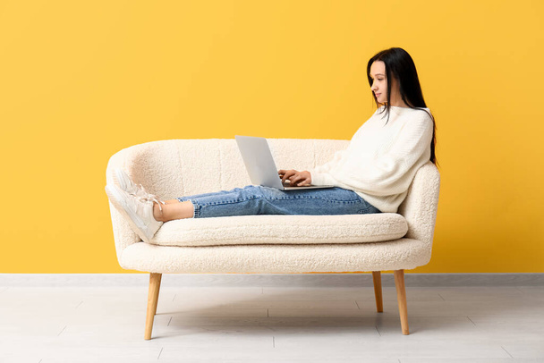 黄色い壁の近くの快適なソファーに横たわっているラップトップを持つ美しい若い女性 - 写真・画像