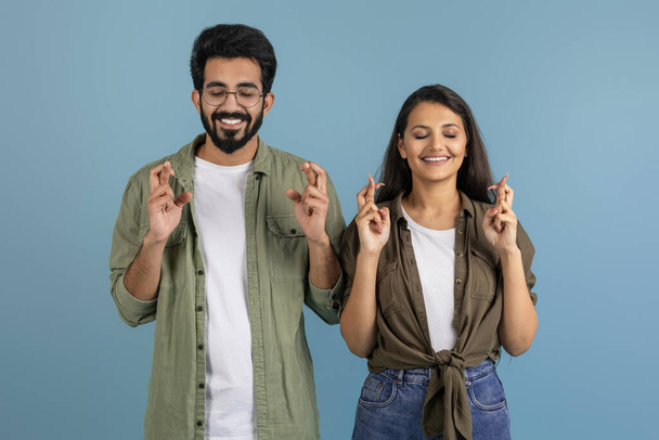 Excited millennial indian couple man and woman wearing casual clothing holding crossed fingers, posing with closed eyes, make wish, cherish hope, isolated on blue studio background - Photo, Image
