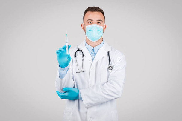 Männlicher Arzt in Schutzmaske und blauen Handschuhen hält selbstbewusst eine medizinische Spritze in der Hand, die die Bereitschaft zur Impfung oder Behandlung repräsentiert - Foto, Bild
