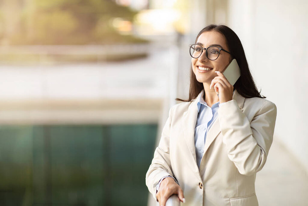 Šťastná inspirovaná mladá běloška v obleku mluví na mobilním telefonu na lehkém kancelářském pozadí. Obchodní komunikace a volání klientovi, práce s gadgetem - Fotografie, Obrázek