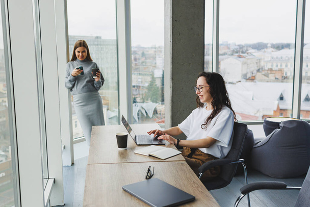 Two young women work in a modern office. Work office space with large windows. Woman manager works at the desk sitting in the office. - Photo, Image