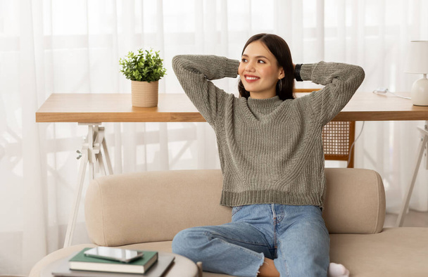 Расслабленная положительная привлекательная брюнетка молодая женщина в повседневной одежде студентка отдыхает дома, сидит на диване с руками за головой, смотрит на пространство для копирования и улыбается - Фото, изображение