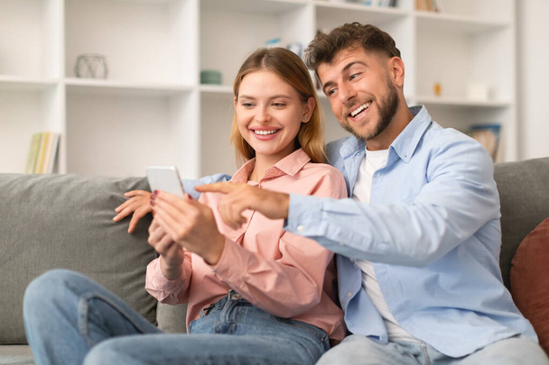 Iloinen tuhatvuotinen pari, jolla on hauskaa matkapuhelimen kanssa, mukavasti sohvalla, selaamassa ja osoittamassa mielenkiintoista internet-sisältöä älypuhelimella, kotona olohuoneessa - Valokuva, kuva