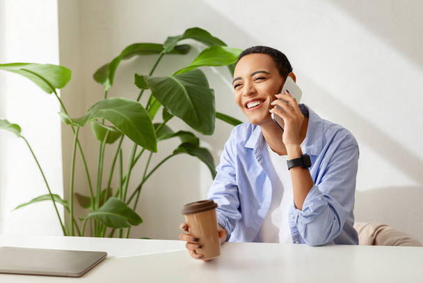 Smiling young lady with short hair, with smartwatch holding coffee cup in hand, talking on mobile phone, with laptop and indoor plants in background. Remote work and communication - Photo, Image