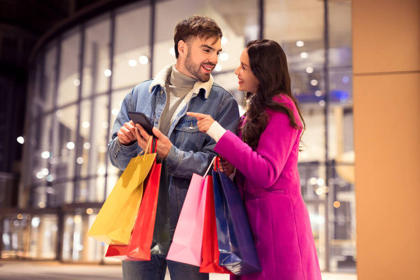 Genç çift kış şenliği sırasında online satış alışverişi için telefonları kontrol ediyor, kış şehrinde akşamları alışveriş torbalarıyla modern alışveriş merkezinin önünde duruyorlar. Noel ve Sevgililer Günü indirimi - Fotoğraf, Görsel