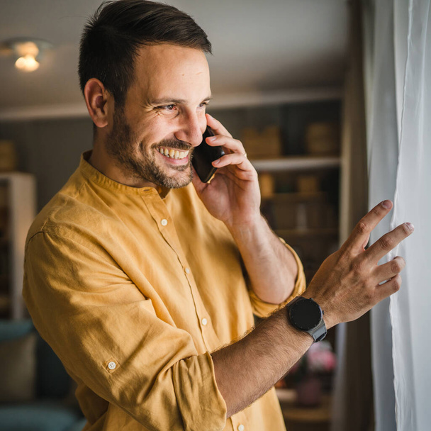 Um homem adulto caucasiano masculino usar smartphone telefone celular para fazer uma chamada falar sorriso feliz em casa usar camisa e óculos espaço cópia pessoa real - Foto, Imagem
