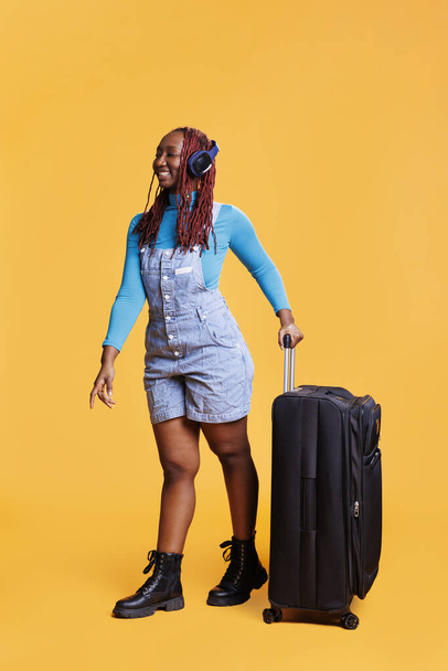 Χαμογελαστή γυναίκα που ακούει μουσική κατά την πτήση, που έχει μεγάλο καρότσι αποσκευών για απόδραση το Σαββατοκύριακο ή διάλειμμα στην πόλη. Ευτυχισμένος νεαρός ενήλικας που φοράει ακουστικά στο ταξίδι των διακοπών, ποζάρει στο στούντιο. - Φωτογραφία, εικόνα