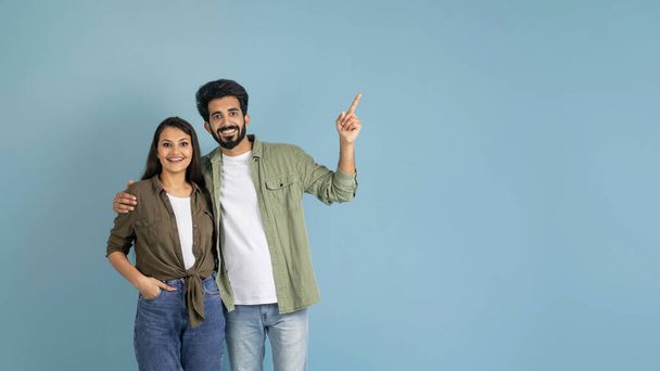 Amazing deal, great offer. Positive young indian couple embracing, showing blank space for text or advertisement isolated on plain blue studio background, panorama - Photo, Image