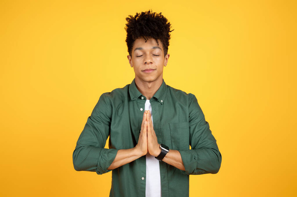 Klidný stylový pohledný mladý černoch, držící se za ruce v namaste gestu, modlící se nebo meditující, izolovaný na žlutém pozadí studia. Úleva od stresu, koncepce bdělosti - Fotografie, Obrázek