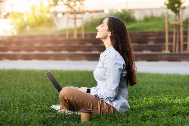 Pozytywny spokojny profesjonalny młody Europejczyk kobieta siedzi z laptopem w trawiastej okolicy na świeżym powietrzu, oczy zamknięte, ciesząc się chwilą spokoju i refleksji. Relaks i detoks cyfrowy w przyrodzie - Zdjęcie, obraz