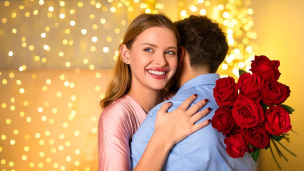 Beaming woman in pink dress embracing man from behind, holds lush bouquet of red roses, with backdrop of golden bokeh lights, panorama with free space - Photo, Image