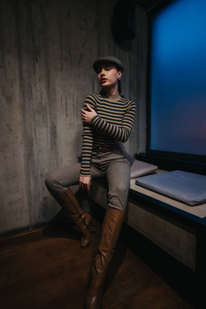 Μια ελκυστική νεαρή γυναίκα γύρω στα 20, να κάθεται μέσα, φορώντας καθημερινά ρούχα και ένα κομψό καπέλο εφημεριδοπώλη. Αποπνέει αυτοπεποίθηση και αίγλη με την κομψή αίσθηση της μόδας. - Φωτογραφία, εικόνα